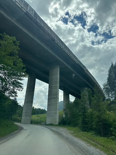 This media file depicts an underpass of a highway bridge in Flachauwinkl, Austria. The perspective is from a roadside, looking upwards at the immense concrete structure and its supporting pillars. Tall, dense green trees and foliage line the road and surround the base of the bridge, indicating a natural, possibly mountainous or forested, environment. The sky above is cloudy with patches of blue visible, suggesting it is daytime with potentially changing weather. The road itself is paved with asphalt and gravel, with a slight sheen indicating it might be damp. The bridge's construction is modern, with clean lines and robust concrete pillars. No people are visible in the image, and there is no visible text. The scene evokes a sense of scale and the impressive engineering required to build such a structure within a natural landscape. The lighting suggests it is daytime, but the overcast sky might temper the brightness. The location cues of dense greenery and the scale of the bridge imply a rural or semi-rural setting within Austria.