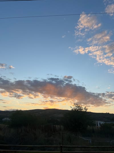 The image depicts a sunset over a rural landscape, likely in or around the village of Djerman, Bulgaria. The sky is a blend of pale blue and warm orange hues, with scattered clouds illuminated by the setting sun. A power line is visible horizontally across the upper portion of the frame. In the midground, rolling hills are covered in dark vegetation. In the foreground, there is a fence and tall grass. A few sparse trees are also present. The overall impression is one of a tranquil, natural setting at dusk.
