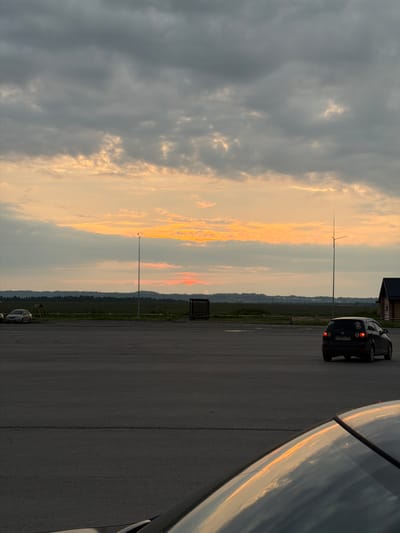 The image shows a sunset scene on what appears to be an airfield or an open, paved area in Ust-Sysolsk, Russia. A dark-colored car is parked on the right side of the frame, facing away from the viewer and towards the horizon. The sky is filled with dramatic clouds, with a band of bright orange and yellow light indicating the setting sun. The rest of the sky is a mix of dark gray and lighter, wispy clouds.

In the background, a thin, tall pole or antenna stands in the distance. Further beyond, a line of trees and hills is visible against the horizon, suggesting a rural or semi-rural setting. The ground is paved, with faint lines suggesting parking spaces or a road. The right side of the image is dominated by the reflection of the sky and clouds on a shiny surface, possibly the hood or roof of another vehicle.

The time of day is clearly sunset, indicated by the vibrant colors in the sky. The weather appears to be partly cloudy. There is no discernible human activity in the scene. The overall mood is peaceful and serene, capturing the beauty of the natural light at dusk.

Visible text on the car's license plate appears to be "P719HT 154".