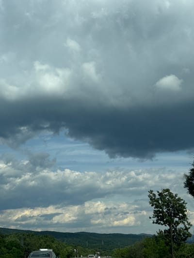 The image depicts a cloudy sky over a hilly, tree-covered landscape. In the foreground, a road with several cars is visible. The sky is filled with a variety of clouds, ranging from light and wispy to dark and heavy. The dark clouds suggest the possibility of rain or a storm. The hills are covered in dense green trees, and the overall impression is of a natural, rural setting. The location is not specified in the image.