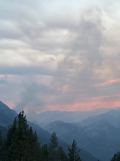 The image depicts a mountainous landscape with a dramatic sky, likely taken from the vicinity of San Juan de Plan, Spain. In the foreground, dark green pine trees are prominent, their tops reaching towards the sky. Beyond the trees, a series of rugged mountains recede into the distance, their peaks shrouded in layers of atmospheric haze. The mountains are rendered in various shades of blue and gray, with each successive range appearing fainter and lighter due to the distance and haze.

The sky is a complex blend of soft grays and pale pinks, suggesting either dawn or dusk. Wisps of clouds drift across the upper portion of the image, with some appearing denser and darker, potentially carrying smoke. In the middle ground, a plume of gray smoke rises from behind one of the mountain ridges, indicating a potential fire or other atmospheric phenomenon. The overall impression is one of natural grandeur and a touch of ominousness due to the visible smoke. The lighting is diffuse, creating soft shadows and a muted color palette across the entire scene.