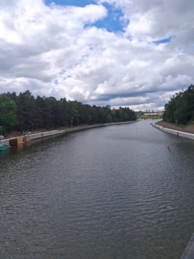 The image depicts a wide canal, identified as the Södertälje Canal in Södertälje, Sweden, under a partly cloudy sky. The sky is dominated by large, dense grey and white stratocumulus clouds, with visible patches of bright blue sky in the upper frame, particularly towards the right. The water in the canal is dark grey-blue, exhibiting ripples and small waves across its surface, indicative of current or wind.

Both banks of the canal are lined with dense foliage, primarily consisting of tall coniferous and deciduous trees, forming a continuous green and dark green barrier. Along the immediate edges of both banks, a modern concrete retaining wall is visible, providing a clear demarcation between the land and the water.

On the left bank, below the treeline, there is an area of apparent ongoing construction or landscaping. Exposed earth is visible, alongside what appears to be a foundation or structural work, and a green tarp-covered object or structure extends into the water. Several streetlights or utility poles are intermittently spaced along the edge of this bank.

On the right bank, a similar concrete retaining wall supports the tree line. A narrow path or cleared area runs along the top of this wall, and streetlights are also present. In the canal itself, two prominent yellow cylindrical marker buoys or poles are partially submerged; one is located in the mid-ground towards the right bank, and another is further down the canal in the center.

In the distant background, beyond the curving canal and the tree lines, several multi-story buildings are discernible, suggesting an urban or suburban environment. No individuals, vehicles, or boats are visible in the immediate foreground or mid-ground. The scene is devoid of direct human activity.