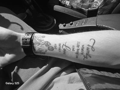 This black and white image depicts a close-up view of a person's left forearm and hand, positioned within the interior of a vehicle, in Novo Delchevo, Bulgaria. The forearm exhibits two tattoos: a stylized illustrative design on the wrist, partially obscured by a dark-colored watch with a prominent buckle, and script text further up the arm reading "Family where life begins and Love never end". The person's left hand is visible at the bottom left, casting a shadow onto a light-colored, textured fabric surface (likely clothing or a seat). To the right of the arm, parts of a car's interior are visible, including a central console with control buttons and a textured finish, and a dark-colored object resembling a gear shifter. A plastic-wrapped item is partially discernible in the background. Strong illumination from an unseen source suggests direct sunlight. The text "Galaxy S25" is visible in the bottom-left corner of the image. No people or actions beyond the arm's presence are depicted.