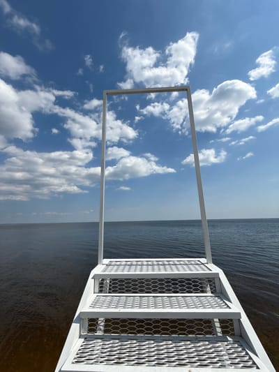 The image displays a white metal structure, likely a pier or waterfront installation, extending into a large body of water under a clear blue sky with scattered white cumulus clouds. This scene is located at a water body near Hlibivka, Kyiv Oblast, Ukraine, specifically on what appears to be the Kyiv Reservoir. The structure is composed of at least three visible steps, each featuring a white, perforated, expanded metal mesh surface for traction. These steps ascend to a prominent, large, open rectangular frame constructed from white metal beams, positioned vertically and centered against the sky. The water in the foreground exhibits a dark brown hue, gradually transitioning to a darker blue-grey as it extends towards the horizon line in the distance. No individuals, vessels, or additional landmasses are visible within the frame. The scene is illuminated by bright daylight, casting minimal shadows.