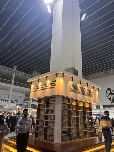 A book fair stand displays books in a grid of wooden shelves, illuminated by spotlights. A person stands near the stand. The structure has an inscription in Arabic. The roof is dark and ribbed. A tall, white pillar extends from the roof towards the stand. People can be seen browsing other stands in the background.