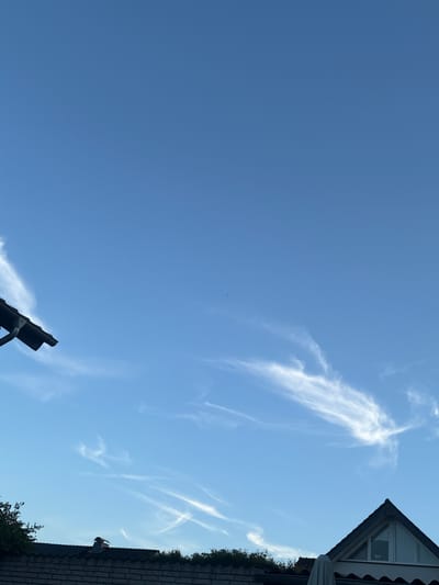 The image depicts a clear blue sky with wispy cirrus clouds. In the foreground, the roofs of houses are visible. On the left, a dark-colored roof with a gutter runs along the top edge of the frame. On the right, the triangular gable of a house with a window is prominent. Vegetation and a chimney are also visible on the rooftops. The scene appears to be an outdoor view from the city of Delbrück, Germany, looking upwards. There are no people or animals present in the image.