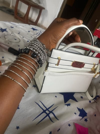 The scene is set indoors, in Odo Ona, Nigeria. The image displays the right arm and hand of an individual with dark skin, gripping the top handle of a small, structured white handbag. The handbag features gold-colored hardware, including a rectangular plaque inscribed "LYN EST. 1991" on the front, decorative straps with gold-colored buckles, and a small open pocket. The individual's wrist and forearm are adorned with numerous bracelets: approximately ten thin, silver-colored bangles embellished with small, reflective stones, several strands of small, spherical silver-colored beads, and at least two larger, spherical black beads. The subjects are positioned on a white fabric surface patterned with blue stars and abstract blue line drawings. Visible on this surface are a silver-colored hoop, a pen with a clear cap and a reddish body, an orange-capped cylindrical object, and a crumpled piece of pink material. The background is out of focus, showing a dark wooden furniture element and an indistinct, darker interior space.
