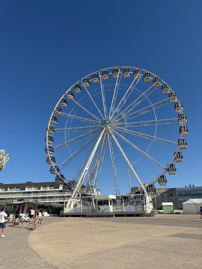 A large white Ferris wheel stands prominently against a clear blue sky in the city of Berck, France. The wheel features numerous enclosed gondolas, many of which are visible, suspended from its outer rim. At the base of the Ferris wheel, a white ticket booth or control center is present, with a sign indicating "POUR VOTRE CONFORT LES CABINES DE LA GRANDE ROUE SONT CLIMATISÉES," meaning "For your comfort, the cabins of the Ferris wheel are air-conditioned."
In the foreground and to the left, a paved area with a curved design separates the open space from a building complex. Several outdoor tables and chairs are set up on the paved area, with a few people visible in the vicinity, including a man in a white t-shirt and dark shorts walking away from the camera. Behind the tables, a low structure resembling a kiosk or food stand is present.
Further back, a long, multi-story building with balconies runs horizontally across the mid-ground, its architecture suggesting it could be a hotel or apartment complex. The Ferris wheel is positioned in front of this building, dominating the scene. To the right of the Ferris wheel's base, a green utility box and a white tent are situated in the open space. Another building, with a more modern facade, is visible behind the tent. The overall environment appears to be an outdoor public space, likely a promenade or plaza, adjacent to a beachfront or waterfront area, given the bright, sunny conditions and the typical seaside amusements suggested by the Ferris wheel.