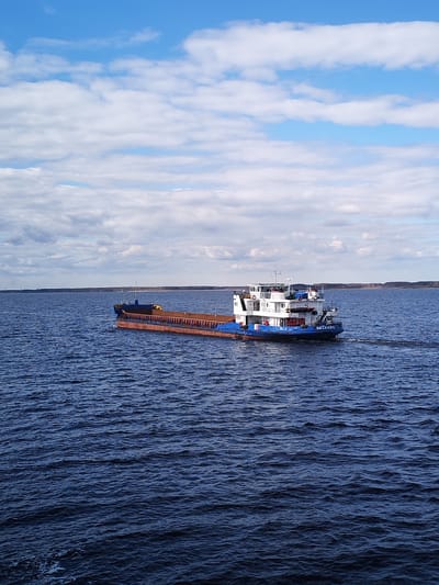 The image captures a cargo ship navigating a large body of water, likely a wide river or lake, possibly near the city of Maksakovo, Russia. The ship is a blue and white tugboat pulling a rust-colored barge with cargo. The body of water has a dark blue hue with visible ripples. The sky is partially cloudy with alternating patches of blue sky and white clouds. A faint line of trees can be seen on the horizon.