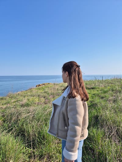 A young woman stands on a grassy cliff overlooking the Black Sea on a sunny day. The location appears to be Sveti Nikola, Bulgaria. The woman is wearing a beige jacket with white fur lining and blue jeans. She has brown hair and is looking down. The grass on the cliff is green and brown, and there are some small purple flowers growing in it. The sky is blue.