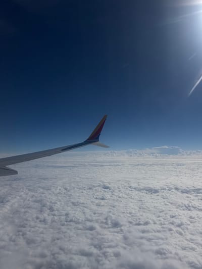 An airplane wing extends into a clear blue sky, capped with a red and yellow tip. The view from the airplane window features a vast sea of fluffy white clouds stretching to the horizon. The sun is bright. The perspective is from above, looking down at the cloud cover.