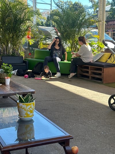 In Bellingen, Australia, two women are seated on an outdoor bench. One woman, with dark hair and glasses, wears a black t-shirt and dark pants, and is touching her hair. The other woman, with her hair in a ponytail, wears a light-colored t-shirt and dark pants and is gesturing with her hands. A baby is crawling on the ground between them, playing with a pink toy and a pink ball. The bench is made of green upholstered furniture and a wooden pallet base, with yellow and silver cushions. Behind the bench is a dark-colored SUV with a license plate ending in "50". To the left of the bench, a black couch is visible, along with potted plants and a dark backpack. In the foreground, a wooden table with a glass top holds a yellow potted plant. Sunlight casts shadows across the concrete ground. A stroller is partially visible on the right side of the image, and a red apple rests on the ground near the table. The environment suggests an outdoor seating area, possibly at a cafe or restaurant.