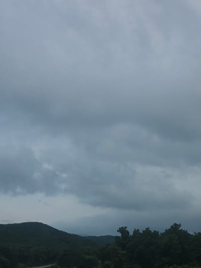 The image depicts a cloudy, overcast sky over a densely forested mountainous landscape. The foreground features the dark silhouettes of numerous trees, with their branches and foliage creating an uneven, textured line against the sky. To the left, a rolling mountain range, also covered in dark green trees, recedes into the distance, its contours softened by the atmospheric conditions. A faint hint of a road can be seen winding through the trees in the lower left portion of the image. The sky is a uniform, muted gray, with subtle variations in cloud density and texture, suggesting a potentially gloomy or rainy day. There are no people or visible structures in the frame. This scene is not specifically identifiable as being in the city of Bolshoy Kamen, Russia, as the image lacks any distinct landmarks or urban elements.