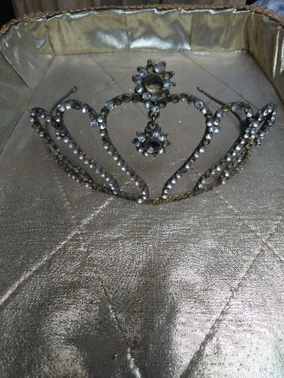 A small, ornate tiara, constructed from tarnished bronze-colored metal and adorned with numerous clear, round rhinestones, is centrally positioned within a shallow, rectangular container. The tiara's design incorporates multiple heart-shaped or arched elements forming its upper edge, each encrusted with small stones. A prominent central decorative element at the apex features a larger starburst pattern with a central stone, from which a smaller, matching starburst pendant is suspended. Several rhinestones are either absent or discolored, and the metal components exhibit visible wear and tarnish. The tiara's left headband arm appears slightly bent. The container's interior is lined with a quilted, lustrous gold-colored fabric, exhibiting a stitched diamond pattern across its surface. The container's upper perimeter is bordered by a slightly darker, textured gold trim. The scene is located in the city of Косшы, Kazakhstan.