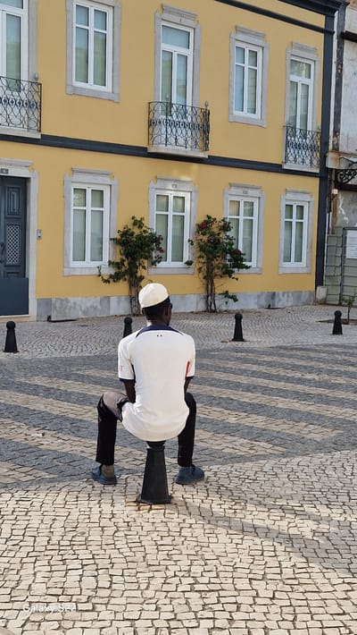 A person wearing a white t-shirt with red and blue accents and black pants is seated on a black bollard facing away from the camera. The person is also wearing a white cap and dark sunglasses. The scene takes place outdoors on a cobblestone plaza in Faro, Portugal, with a yellow building in the background featuring multiple windows and balconies. The cobblestones are arranged in a pattern of light and dark gray stripes.