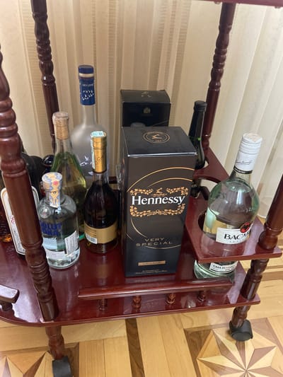 The image shows a wooden wine rack filled with various alcoholic beverages. The rack is brown with a glossy finish and has multiple tiers to hold the bottles.
The top tier contains a bottle of Bacardi rum and another unidentified green bottle. The second tier has a bottle of Hennessy Very Special cognac in its box and another dark bottle. The third tier contains a bottle of Grey Goose vodka, an unidentified green bottle with a cork, and a bottle of Absolut Vodka. The bottom tier has two unidentified bottles.
The background consists of a white curtain and a wooden floor with a star pattern.
Based on the presence of various international alcoholic brands, it suggests the location could be a home, bar, or retail store. However, without additional information, it's impossible to confirm the location to Akhalkalaki, Georgia.