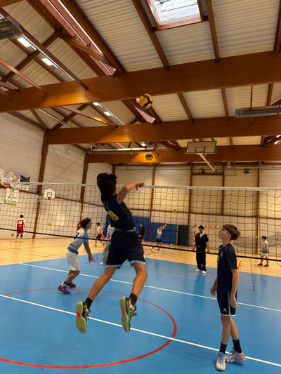 The image captures an active indoor volleyball scene within a gymnasium located in Sennecey-le-Grand, France. The environment features a blue court with distinct white and red markings, a black volleyball net spanning the width of the court, and a high ceiling composed of exposed wooden beams, white ribbed panels, and visible skylights. Overhead lighting fixtures are also present. A basketball hoop with a backboard is discernible in the background.
In the foreground, a young male with dark hair, wearing a dark blue short-sleeved shirt with yellow detailing and dark blue shorts with light blue trim, is captured mid-jump with his right arm extended upwards, poised to strike a yellow and blue volleyball suspended in the air above him. He wears bright green and black athletic shoes. To his right, another young male in a dark blue short-sleeved shirt (with an 'Errea' logo visible) and dark blue shorts stands, looking upwards towards the airborne ball and the jumping player. He wears light blue and white athletic shoes.
Further left and slightly behind the main jumping player, a third young male with light hair, dressed in a light blue and white short-sleeved shirt and white shorts, stands near the net, facing right. Multiple other individuals, primarily young males in various athletic attire, are visible in the background, dispersed across the court and near the net, suggesting a training session or game in progress. One player in the far-left background wears a red jersey with the number '63'. Another individual in the mid-background wears a black tracksuit and appears to be observing the activity.