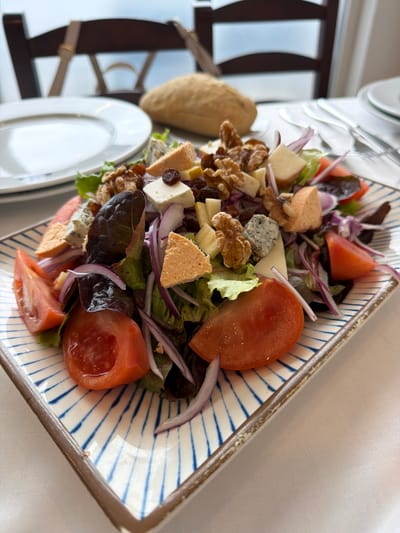 An overhead, slightly angled close-up view depicts a prepared salad centered on a rectangular white plate with blue radial stripes and a brown rim, positioned on a white tablecloth. The scene is set indoors, likely a restaurant, in Colunga, Spain.

The salad consists of a base of mixed green and red leaf lettuce. Distributed throughout are multiple roughly quartered red tomatoes, thinly sliced red onions, and various cheese types: irregular chunks of blue cheese, cubes of a pale yellow firm cheese, triangular pieces of a light orange cheese, and cubes of a white, possibly crumbly, cheese. Also present are whole and partial walnut pieces and dark raisins. A light dressing is visible on the ingredients.

In the blurred background, a rustic-style bread loaf is visible on the tablecloth behind the salad. To the left of the bread, a plain white round dinner plate is stacked. To the right, silverware including a fork and knife rests on a white napkin. The back of a dark brown wooden chair is visible, with a light-colored strap draped over it. Beyond the chair, a large, out-of-focus window reveals a bright, diffuse outdoor view.