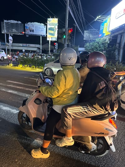 This media file captures a nighttime scene on a street in Denpasar Barat, Indonesia, likely during a traffic light stop. Two individuals are seated on a scooter. The person in front wears a yellow jacket and a silver helmet, while the person behind them is dressed in a dark hoodie and a maroon helmet. They appear to be riding together, with the person in front operating the scooter.
Several advertisements are visible in the background. One prominent billboard displays "MODERN KRETEK IN Pop-Up PACK" and "DUNHILL." Another banner reads "SEKOLAH DIAN HARAPAN GLOBAL BALI" with an opening date of "JULY 2017," suggesting an educational institution. There are also traffic lights, indicating an intersection. The street is asphalt, and the lighting suggests it is nighttime. The overall atmosphere is that of a typical urban street scene in Southeast Asia at night, with the presence of traffic and illuminated advertisements. Visible text includes brand names and information about a school.