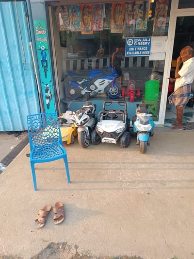 The scene depicts a street-level retail storefront in Anantapur, India, during daytime. A partially raised blue corrugated metal shutter is on the far left. Immediately adjacent is a green vertical sign for "Enviro SRINIVASA E-BIKE" featuring scooter images. The central section is a large glass window displaying a blue and white toy motorcycle, a red tricycle, and a green barrel inside. Promotional signage on the glass includes "BAJAJ FINSERV EMI FINANCE AVAILABLE HERE" and a "KIDS TOYS" sign with a contact number. Four children's ride-on vehicles are arranged on the concrete sidewalk in front of the store: a yellow car, a silver motorcycle, a white all-terrain vehicle (ATV) style vehicle, and a blue motorcycle. A bright blue plastic chair with a patterned backrest is positioned on the left side of the sidewalk. A pair of brown leather sandals lies on the concrete ground in front of the chair. An elderly man stands on the right, barefoot, wearing a white shirt and a plaid lungi. He is touching his right temple with his right hand. A reflection of another individual in a dark shirt and shorts is visible inside the store, reflected in the glass window. The ground is a light-colored concrete surface.