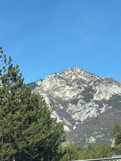 The image displays a majestic natural landscape featuring a prominent mountain face, a clear blue sky, and dense green foliage. The original image appears to be rotated 90 degrees clockwise; when viewed in its intended vertical orientation (rotated counter-clockwise), the scene becomes clearer.
In this vertical orientation:
On the right side of the frame, a large, imposing rock face of a mountain dominates. This rock face is a mix of light grey, white, and beige tones, with intricate textures, shadows, and crevices suggesting its ancient, weathered form. A notable feature of this mountain is the striking resemblance of its left profile to a human face. This natural formation clearly defines a forehead, a prominent nose, and a distinct chin, all looking towards the left into the clear sky.
The left side of the image is filled with a vast expanse of bright, clear blue sky, devoid of clouds, indicating fair and sunny weather.
In the foreground, particularly across the bottom of the image and extending up the lower left side of the mountain, is a dense mass of dark green trees. These appear to be coniferous, with their thick foliage creating a rich, textured foreground against the rugged mountain and bright sky. Further up the mountain, beyond the main rock face, slopes are covered in lighter green vegetation.
Minimal man-made elements are present:
Towards the top right portion of the mountain, against a backdrop of green foliage, a thin white pole with a small, dark, rectangular object (possibly a solar panel or sensor) attached is visible. In the very top right corner, a segment of a white vertical structure, possibly a pole or part of a distant building, can be seen.
The time of day is clearly daylight, likely mid-day, given the bright illumination and clear sky. There is no visible text in the image. The most distinctive aspect of the image is the natural pareidolia, where the mountain's profile strikingly resembles a human face.