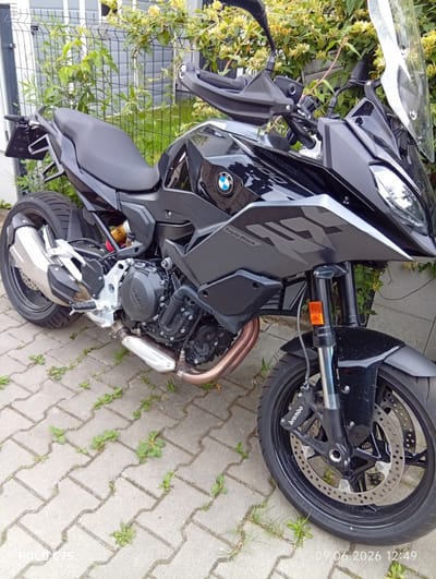 A black BMW F900XR motorcycle is parked on a paved patio with green plants growing between the pavers, in front of a gray fence and building. The motorcycle is angled towards the viewer's left, with its front wheel facing forward. The bike features a black seat, black fairing with a silver stripe, a clear windscreen, and a silver exhaust pipe. The BMW logo is visible on the fairing. The pavement appears to be made of interlocking concrete blocks. The background shows a chain-link fence with lush green foliage and yellow flowers. The scene suggests an outdoor setting, possibly a residential area or a parking lot. The timestamp "09.06.2026 12:49" is present in the bottom right corner of the image.