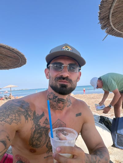This is a close-up, portrait-style photograph taken from a low angle, looking up at a man on a sunny beach. The man is wearing a grey baseball cap and sunglasses, and his chest and arms are visible, covered in various tattoos. He is holding a clear plastic cup with a drink and a blue straw.
In the background, a beach is visible with other people, beach umbrellas, and the sea. The sky is a clear, bright blue, suggesting it is daytime with good weather. The sandy beach is visible in the middle ground, and further out, the water stretches to the horizon.
There are several people in the background. One man, wearing a green t-shirt and a white baseball cap, is bent over and holding a small booklet. Another person, with tanned arms, is seen from the shoulder up, also on the beach. In the distance, several small figures and beach umbrellas are scattered along the shoreline. Two thatched beach umbrellas are prominently featured in the foreground and bottom left of the image, framing the shot.
The tattoos on the man's body are detailed and cover a significant portion of his chest and upper arm. Visible text includes "MEMORIA" tattooed vertically on his chest. The overall mood seems relaxed and vacation-oriented, typical of a day at the beach. The orientation of the photo is rotated.