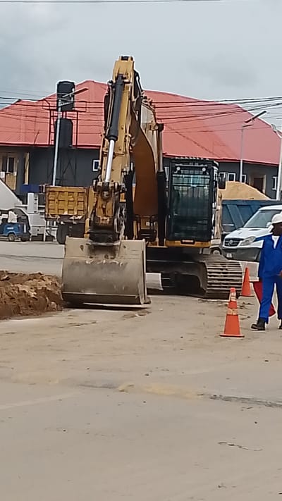 An excavator with a large bucket is positioned in a dirt area. Behind it, a yellow dump truck is parked. A white van is visible to the right of the excavator. Two orange traffic cones are placed on the ground in front of the van and excavator. A person wearing a blue uniform and white hard hat is standing to the right of the traffic cones, facing towards the right of the image. The background includes a red-roofed building with dark walls and some overhead wires. The ground is a mix of dirt and paved surfaces. The overall scene depicts a construction or road work environment.