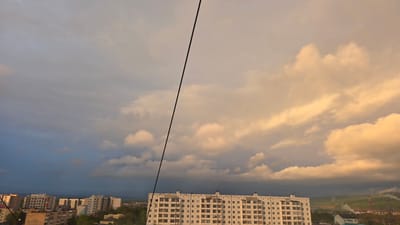 The image captures a cityscape of Lensk, Russia, under a sky with dramatic cloud formations. A large, white apartment building dominates the lower center, appearing to be a typical Soviet-era design with many windows. To the left of this building and farther in the distance are several other residential buildings, suggesting an urban setting. In the background, there's evidence of industrial activity, including smoking chimneys, indicating the presence of factories or power plants. The sky is divided, with a darker, stormy section on the left and a golden, sunlit area on the right, giving a sense of contrasting weather conditions. A black wire is visible diagonally across the sky, likely an electrical or communication line. No people are visible. The scene is static, with the primary focus on the buildings and the sky's atmospheric conditions.