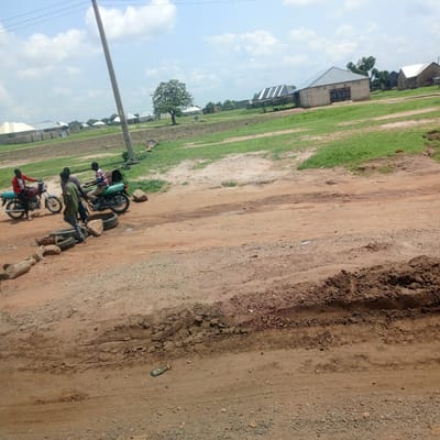 A group of young men are gathered around motorcycles on a dusty, unpaved road in a rural African village. The scene is set in Mutum Biyu, Nigeria, under a bright, cloudy sky, suggesting it's daytime. The ground is a mix of dry dirt, gravel, and some patches of green grass. In the background, simple houses with corrugated metal roofs are visible, along with a single large tree.
One man in a red jacket is sitting on a red motorcycle, while another person in a striped shirt is on a green motorcycle. A third man, wearing a green shirt, stands near the motorcycles, interacting with the group. Several old tires and rocks are scattered on the ground near the men. The overall atmosphere appears casual, with the men seemingly engaged in conversation or preparing for a journey. The presence of motorcycles and the unpaved road indicate a mode of transportation common in such areas. The buildings in the background are modest, typical of rural settlements. There are no visible signs or text in the image.