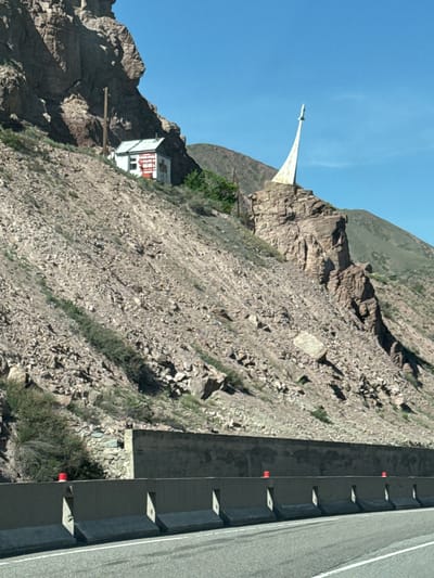 A wide-angle, eye-level shot captures a rugged mountain landscape under a clear blue sky, likely in the Krasnyy Most region of Kyrgyzstan. A paved road, possibly a highway, runs along the base of a steep, rocky incline in the foreground. The road features a substantial concrete barrier with regularly spaced support blocks and reflective red markers separating it from the mountain.

Dominating the scene is a very steep, arid, and rocky mountain slope, heavily covered in light-colored stone debris. Sparse patches of green bushes and grass cling to the rough terrain. High on a prominent rocky peak, a striking white monument stands silhouetted against the bright sky. This monument features a broad, sweeping base that tapers upwards into a slender, pointed spire, resembling a stylized rocket or a soaring wing.

Further down the slope and to the right of the monument, a small, white, flat-roofed building with red accents (possibly trim or a door) is nestled into the mountainside. A wooden plank or bridge extends from the building, potentially serving as an access point or viewpoint. In the background, beyond the immediate rocky slope, gentler, green-hued mountain ranges rise towards the horizon. The bright, even lighting suggests it is midday or early afternoon, with clear weather conditions. No people or visible text are present in the image.