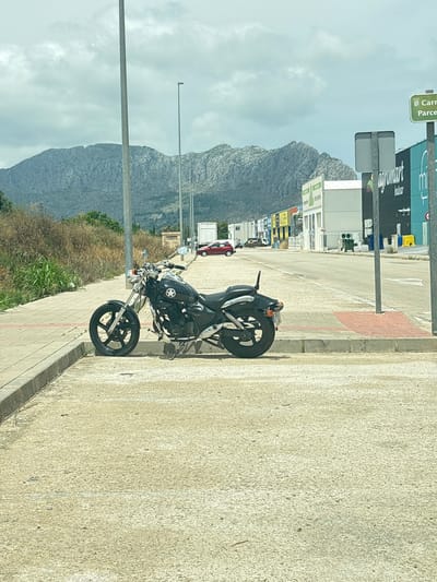 A black motorcycle is parked on a paved area next to a sidewalk, with a street and commercial buildings in the background. In the distance, a mountain range is visible under a cloudy sky. A green sign with white text that reads "iii Carrefour Parc" is present on the right side of the image. The scene appears to be located in the city of Ondara, Spain. The motorcycle is a cruiser-style bike with chrome accents. The commercial buildings include signs for "agromart" and what appears to be a supermarket. A red car is parked further down the street. The environment is an urban or semi-urban setting with vegetation along the sidewalk and a dry, scrubby area behind it.