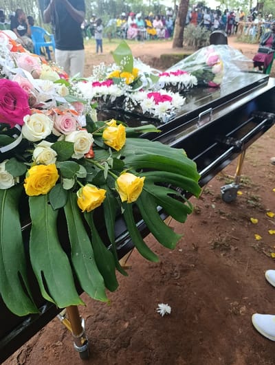 The scene depicts an outdoor funeral gathering in Muthetheni ward, Kenya. A dark-colored, possibly black, casket is positioned on a metallic wheeled stand in the foreground. The casket is extensively adorned with floral arrangements. On the left side of the casket, a large arrangement features pink, white, cream, and yellow roses, along with white filler flowers and large green Monstera leaves. Further along the casket, additional bouquets and wreaths composed of white chrysanthemums and pink/red roses are visible, with one bouquet wrapped in clear plastic. The ground is reddish-brown soil, with scattered white and yellow flower petals. In the lower right foreground, the feet of an individual wearing white sneakers with yellow laces are visible. In the background, a crowd of approximately 50-70 individuals, including adults and children, are gathered, with many seated on plastic chairs. A man in a black shirt and light-colored trousers stands in the mid-distance, holding a device that appears to be for recording. Trees and natural foliage form the backdrop of the gathering.