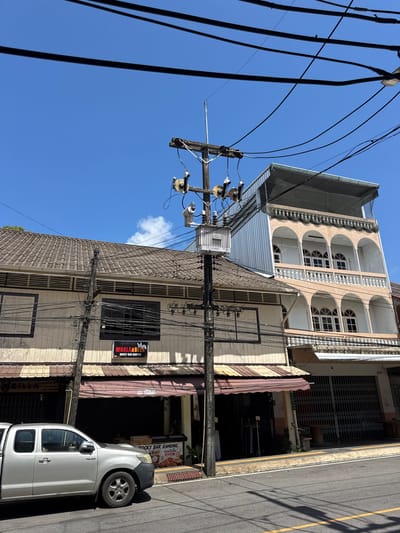 This is a daytime, eye-level, full shot of a street scene in Ranong, Thailand. A silver Toyota Hilux pickup truck is parked on the right side of the road. The truck is facing left and is partially obscuring a building. The building is a two-story structure with a light-colored facade and a tiled roof. Above the first floor, there are several windows and louvers. A dark awning extends over the ground floor, beneath which are closed shutters. A sign above the awning reads "MHALA&BEER ROCKY BAR RANONG". A telephone pole with numerous wires and a transformer is positioned in front of the building. To the right of this building is a taller, multi-story building with a pale pink facade, arched windows on multiple levels, and ornate white railings. The sky is clear and blue with a few white clouds. The road in the foreground is asphalt with a yellow line marking the edge.