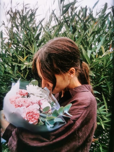 A young woman with long, dark brown hair pulled into a ponytail, wearing a dark reddish-brown collared shirt, is standing outdoors amidst lush green foliage. She is holding a bouquet of pink and white flowers, with green leaves, close to her face as if smelling them. The background is filled with dense green leaves and stems, suggesting a natural, possibly garden or park, setting. There is no specific indication of the city of Serov, Russia within the image itself. The lighting appears to be natural, possibly overcast.