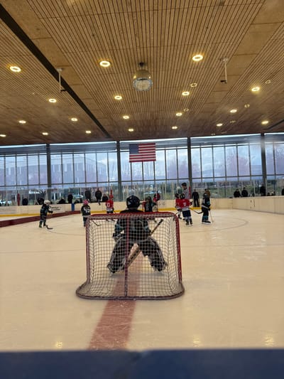 An indoor ice hockey rink shows a game in progress. Players wearing hockey gear are visible near the net, guarded by a goalie. A line of spectators stands against a backdrop of windows. The American flag is hung against the window. The roof has a light wood paneling with round lights installed in rows. A clock hangs on the wall as well.