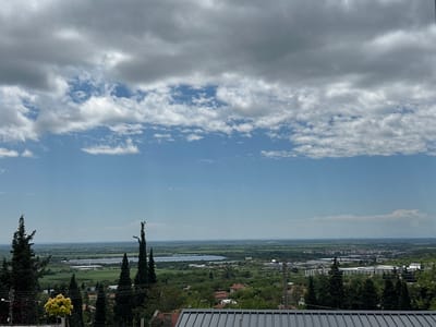 The photograph, taken in Hrishteni, Bulgaria, presents a wide-angle, elevated view of the landscape under a partly cloudy sky. The upper portion of the image is dominated by a dense cloud cover with patches of blue sky visible in between.
In the middle ground is a panoramic expanse of rural and urban areas. Agricultural fields, clusters of trees, and scattered residential buildings comprise the rural component. A large solar panel array is visible in the distance, indicating an industrial or renewable energy installation. Closer to the right side of the image, an urban area is evident, with buildings of varying sizes and designs.
The foreground includes elements from the vantage point, likely a building rooftop. Trees, including cypress-like varieties, frame the lower edges of the image. A low-rise building with a corrugated metal roof runs across the bottom of the frame. Telephone poles and wires are visible among the trees in the foreground, suggesting the presence of infrastructure.