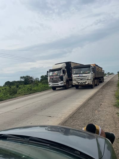 This media file depicts a road scene in Oduape, Nigeria. The primary subjects are several large trucks, parked or moving slowly in a convoy on a paved road. The trucks appear to be hauling cargo, with some carrying what looks like lumber or similar materials. One truck in the foreground is white with "GPC" branding and the number "694" visible on its side. Another truck behind it is black. The road is flanked by a rougher shoulder and a grassy, tree-lined area. In the foreground, the corner of a dark-colored vehicle, likely a car, is visible, indicating that the photo is taken from inside or next to this vehicle. A person wearing a yellow and black striped shirt and black pants is partially visible near the car, leaning down. The sky is overcast with clouds, suggesting a potentially humid or rainy day. The overall impression is that of a truck convoy on a highway in a rural or semi-rural setting.
