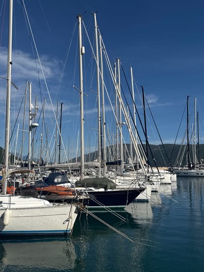The image depicts a marina in Meljine, Montenegro, filled with yachts. The composition focuses on the masts of numerous sailboats docked side-by-side in the harbor. The masts are predominantly white and reach towards a clear, blue sky with scattered cirrus clouds. The masts create a dense network of vertical lines, contrasting with the blue sky and water. The hulls of the boats are visible in the foreground and middle ground, with varying colors and details. The water reflects the sky and the hulls of the boats, creating a mirror-like effect. In the background, there is a hilly landscape covered with vegetation.