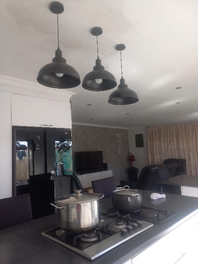 A kitchen scene features three black dome pendant lights hanging against a white wall. Below, a kitchen counter with a gas stove houses two pots. In the background, a black refrigerator stands beside a television. Furniture is visible in the adjoining room, including a black recliner.