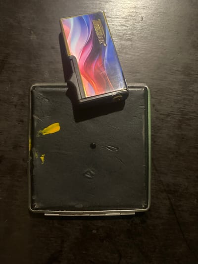The image shows a dark brown, textured surface, likely a wooden table. Placed on the surface are two objects: a black cigarette case and a lighter. The cigarette case is rectangular with a metal frame and a black, textured surface. There are smudges of yellow paint on the cigarette case. The lighter is positioned on top of the cigarette case and has a colorful, abstract design featuring swirls of pink, blue, and white. The lighter also has some text on it that appears to be in a script font. The overall lighting in the image is dim, suggesting it was taken indoors in low light conditions. There are no people visible in the image. The scene is consistent with an indoor setting, potentially a home or workspace. No specific landmarks or street views are visible, and therefore, it is not possible to determine the location within Tyvriv, Ukraine, based solely on this image.