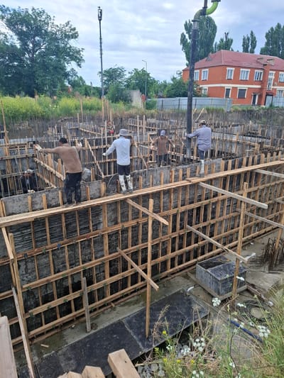 The image depicts a construction site in Argun, Russia, likely a foundation pour for a building. Several construction workers are present, some wearing hard hats and safety vests. One worker is operating a concrete pump, directing a large pipe into the foundation forms. Others are supervising or preparing the site. The foundation area is heavily reinforced with rebar and surrounded by wooden formwork. The surrounding environment includes a cloudy sky, trees, a fence, and a brick building in the background. There are also construction materials and equipment scattered around the site.