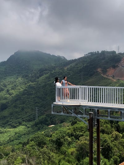 In Nam Hải, Vietnam, two women stand on a metal observation deck overlooking a lush, mountainous landscape. The woman on the left wears a white top and dark shorts with a black hat. The woman on the right is in a pink dress and a denim vest, also wearing a hat. The observation deck, with its white railing, extends outwards over a steep, green hillside. Power lines and a transmission tower are visible in the distance, amidst the dense vegetation and rolling hills. The sky is overcast, with clouds partially obscuring the peak of a large mountain in the background. The scene suggests a tourist attraction offering panoramic views of the natural scenery.