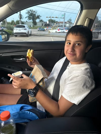 A young boy wearing a white t-shirt and black shorts sits in the passenger seat of a vehicle, looking at the camera and smiling. He holds a sandwich in his left hand and a small container of dip in his right. A brown paper bag is also in his hands. A black wristwatch is on his left wrist. The interior of the car is visible, including a black seat, a seatbelt, and a car door. Outside the car window, cars, trees, buildings, and traffic signals are visible, suggesting an urban or suburban setting. The sky is cloudy. The location is identified as Conroe, United States of America.