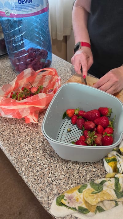 A top-down shot shows a person preparing strawberries in a kitchen setting. The person, wearing a black shirt and a red Apple watch, is using a knife to cut strawberries on a wooden cutting board. A light blue colander filled with ripe, red strawberries sits on a granite countertop. Beside the colander is a clear plastic bag with red stripes, partially filled with strawberry stems. In the lower left corner, a large blue plastic bottle labeled "Devin" is visible, containing water and some strawberries inside. A patterned kitchen towel with lemons is visible at the top right corner of the frame.