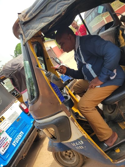 The scene is purportedly set in Candelaria, Colombia, though visual evidence contradicts this assertion. An outdoor shot details a male driver positioned within a three-wheeled auto-rickshaw. The vehicle features a brown, yellow, and blue color scheme with a dark canopy, showing signs of wear, rust, and dirt. The driver, an adult male with dark skin and a shaved head, wears a dark blue long-sleeved shirt with white stripes on the shoulders, tan trousers, and dark slide sandals. He holds a red smartphone in his right hand, directing his gaze downward, with his lips slightly parted. A blue plastic storage basket is visible near the steering column. Partially legible text on the auto-rickshaw's blue lower section reads "NAU NO.". In the background, a second male subject, bearded and wearing a red patterned shirt, is partially visible, looking forward. To the left, a second blue three-wheeled auto-rickshaw is parked, displaying "STUDENTS" in prominent white lettering, alongside "NIZIK TRANSPORT SERVICES 2024" and "NAU". The ground beneath the vehicles is unpaved earth. Distant background elements include sparse vegetation and a building featuring a reddish-brown roofline. The text "NAU" and "NIZIK TRANSPORT SERVICES" visible on the secondary vehicle are strong indicators of a location associated with Nnamdi Azikiwe University in Nigeria, not Candelaria, Colombia.