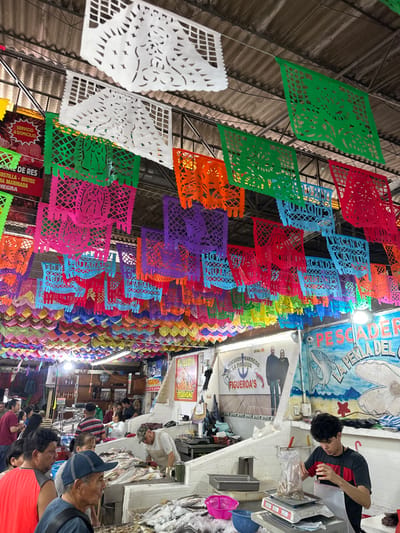 The image shows an indoor market with colorful, intricately cut paper decorations hanging overhead. People are browsing stalls selling fish and other goods. Signs advertise "Pescaderia del" and "Figueroa's." Various colors such as white, green, orange, red, pink, purple, and blue can be seen on the decorations. A couple of people are at a stall selling fish and weighing the products.