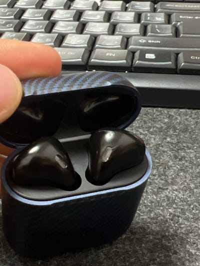 A close-up shot captures a pair of black wireless earbuds inside their charging case, which has a blue carbon fiber pattern. The earbuds are glossy and molded into an ergonomic shape. In the background, a keyboard is partially visible, with keys displaying Cyrillic characters, indicating its use in a region where this alphabet is prevalent. A person's thumb and index finger are seen holding the charging case, suggesting interaction with the device. The scene appears to be indoors, with the textured surface beneath the case hinting at a desk or table. The overall lighting is natural, highlighting the shiny texture of the earbuds. The location, Kadamjay, Kyrgyzstan, is a mining city known for its antimony production, and while not directly depicted, the presence of a keyboard and electronics suggests a modern setting within the city.