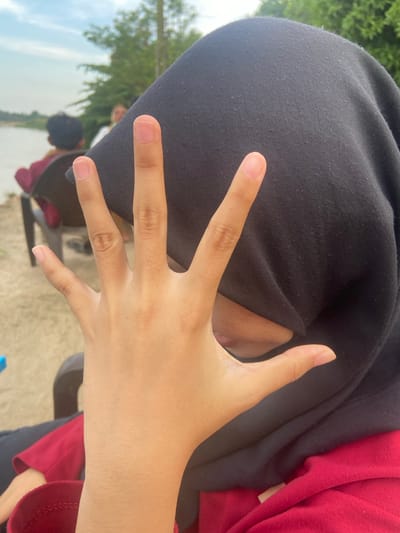 The media file shows a close-up of a person's hand with their fingers spread out, partially covering their face, which is obscured by a black head covering. The person is wearing a red long-sleeved shirt. In the background, out of focus, trees and a body of water are visible, suggesting an outdoor setting. There are other people in the background, one of whom appears to be wearing a black head covering and a maroon shirt. The scene seems to be taking place during the daytime, with natural lighting. The image is taken from a slightly downward angle, looking towards the person's hand. The focus is on the hand and the black head covering. No text is visible in the media file.