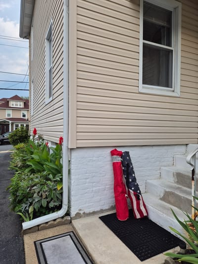 The image shows the exterior of a two-story house in South River, United States of America. The house has light beige vinyl siding and white trim around its windows. A white downspout runs vertically along the corner of the house, feeding into a horizontal drainpipe. In front of the house, a row of green plants with red flowers, possibly canna lilies, are visible. A black car is parked on the asphalt driveway to the left of the plants.

In the foreground, two rolled-up items lean against the white-painted brick foundation of the house. One is predominantly red with a silver reflective stripe, and the other is an American flag with red and white stripes and a field of white stars on a blue background. Both are standing upright on a black rubber doormat. Concrete steps lead up to a doorway, with a rusty metal handrail on the right side of the steps. A gray doormat is situated on the concrete sidewalk to the left of the steps. The sky above is partly cloudy.